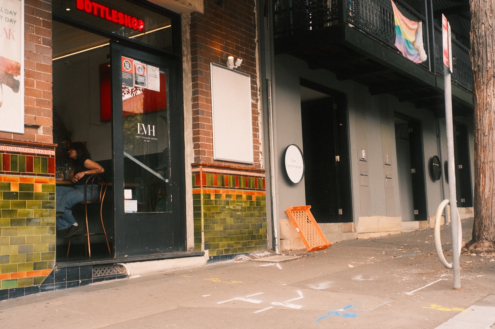 A figure sits at a table inside a bottle shop, seen through the glass; chalk marks cover the pavement outside.